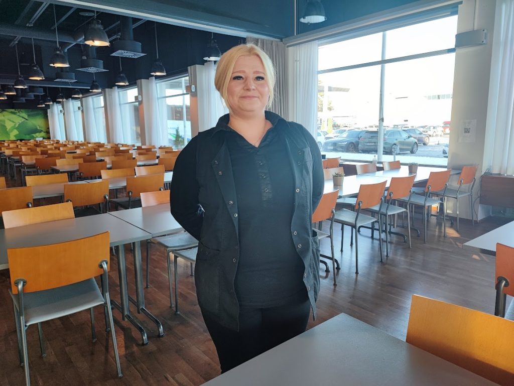Nainen seisoo työpaikan ruokalassa ja hymyille.
